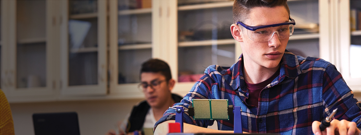 A Student wearing eye-glasses is working in a technology lab. One more student in the background too is working in the lab.